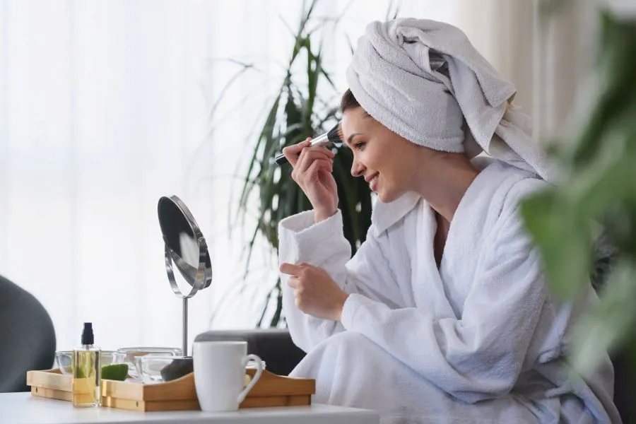 A woman sitting in a robe surrounded by plants putting makeup on.